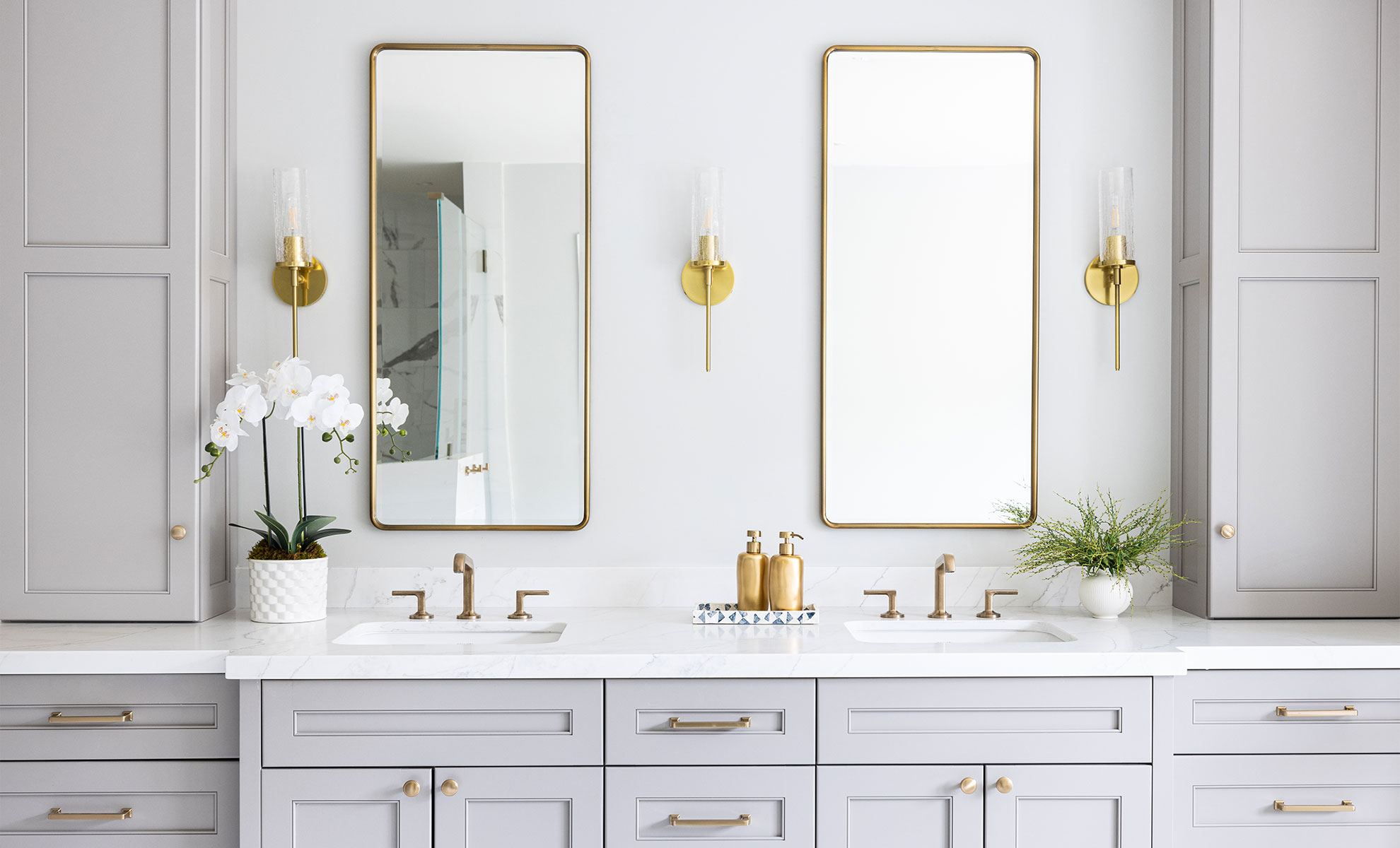 Elegant bathroom vanity with gray cabinets, gold accents, double mirrors, and a white quartz countertop, decorated with orchids and greenery.